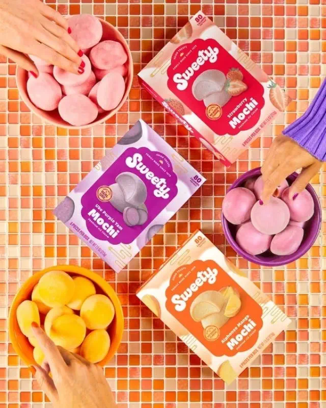 People’s hands reaching for different flavors of packaged mochi on a tiled surface. Boxes include "Sweety," "Mochi," and "Ube Purple Yam Mochi" brands