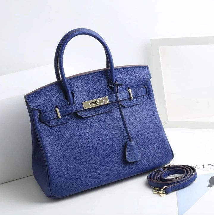 A structured handbag with a top handle and detachable strap is displayed on a surface, showcasing its clasp detail and textured surface