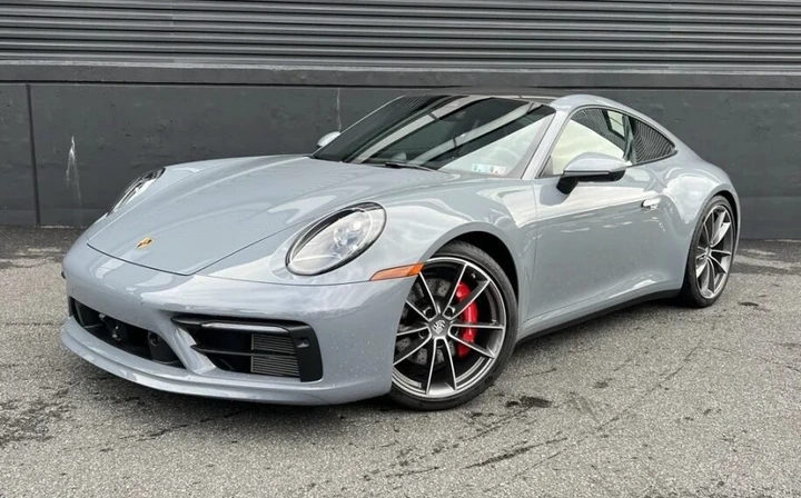 A Porsche 911 Carrera S is parked on a gray asphalt surface in front of a striped black and gray wall.