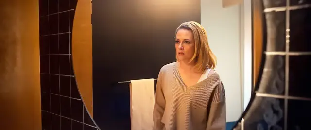 Person with shoulder-length hair looks into a bathroom mirror, wearing a casual oversized sweater