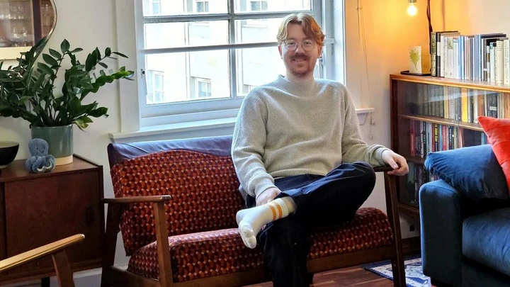 Matthew McCallum Matthew McCallum sits on a bright orange and red subway seat converted into a couch with wooden armrests and legs. He is in a living room by the window. He has ginger hair and is wearing a grey jumper, navy trousers and white socks.