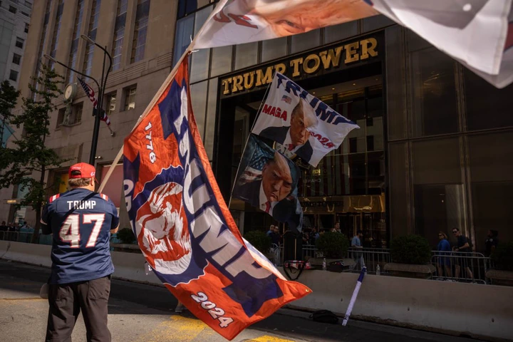 Person wearing a &quot;Trump 47&quot; jersey stands outside Trump Tower holding flags with pro-Trump messages and images