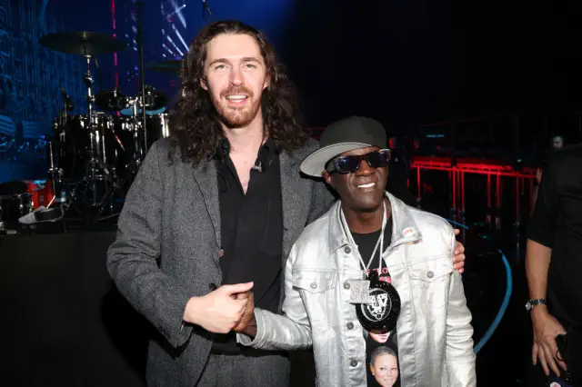 Hozier in a suit and Flavor Flav in a shiny jacket, hat, and sunglasses posing together at an event. Flavor Flav comes to Hozier&#x27;s shoulder