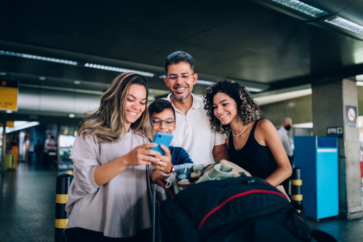 Family of four happily gathers around a smartphone, taking a selfie at an airport terminal, with luggage in tow