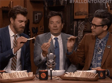 Three men in suits on a talk show eating chips, seated at a table with microphones