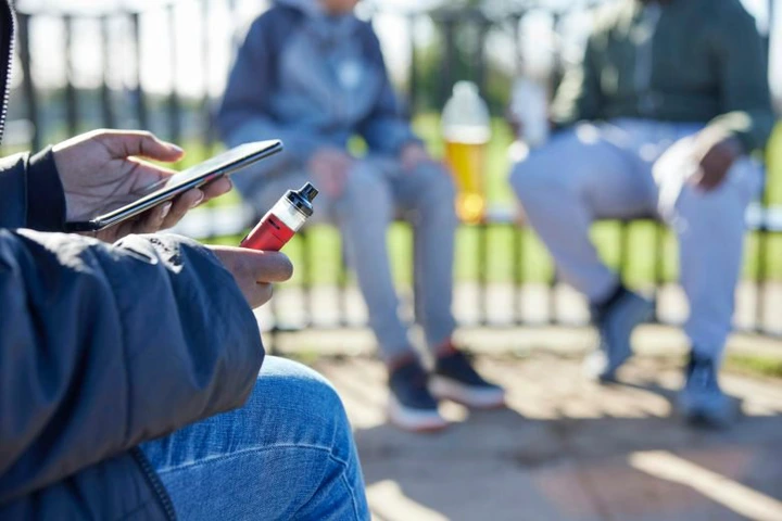 Close-up of teenagers sitting on benches, one holding a vape cartridge and cell phone in foreground.