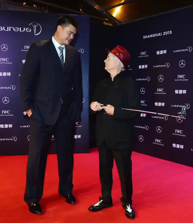 on a red carpet, Yao in a suit and Bill in a formal outfit with a bright hat. They look at each other, engaged in conversation. Bill doesn&#x27;t reach Yao&#x27;s shoulder