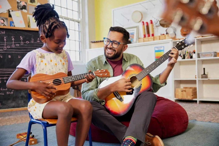 A joyful male teacher is helping an African child learn to play a musical instrument in a classroom setting. The cheerful teacher strums an acoustic guitar and sings during the music lesson, while a girl plays the ukulele beside him.
