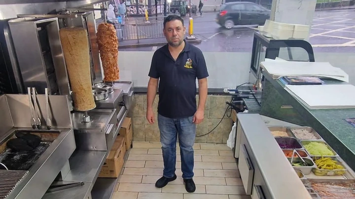Yasin Bilic A man in a blue short sleeve top and jeans stands in a kebab shop behind the counter with a street in the background.