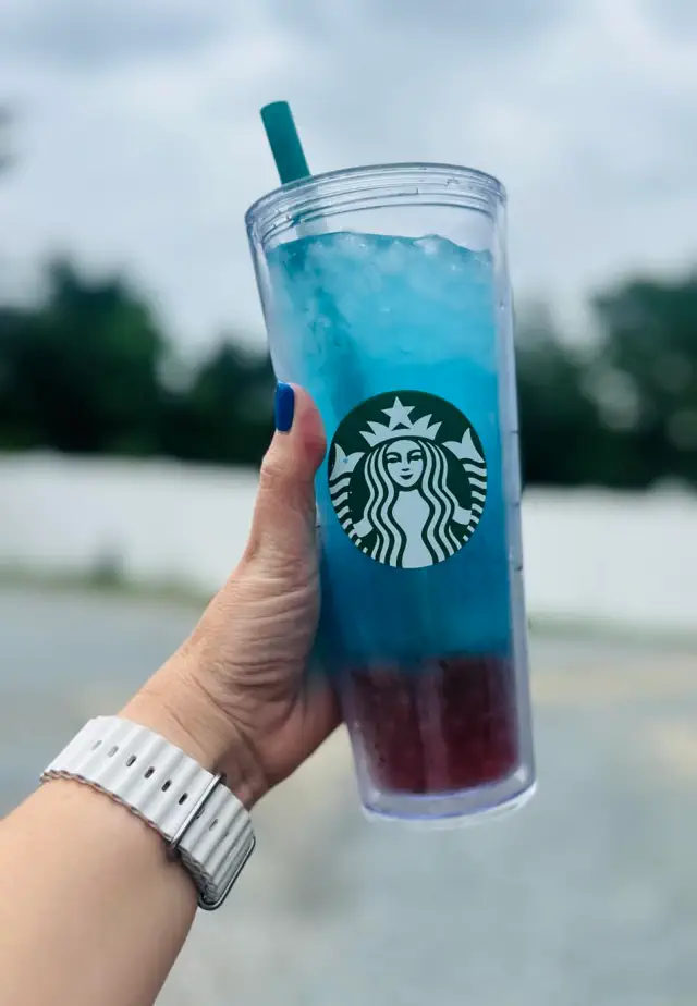 A hand with a smartwatch holds a Starbucks tumbler with a layered iced drink outside against a blurry background