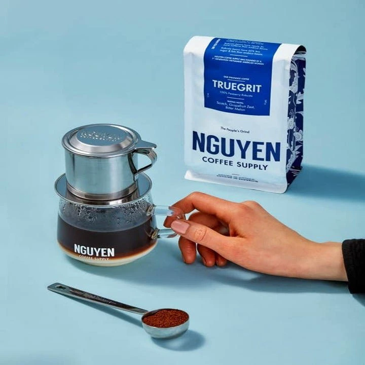 A hand holds a glass of coffee brewed using a stainless steel trfilter. Beside it is a bag of Nguyen Coffee Supply's Truegrit coffee. A teaspoon of coffee grounds is also shown