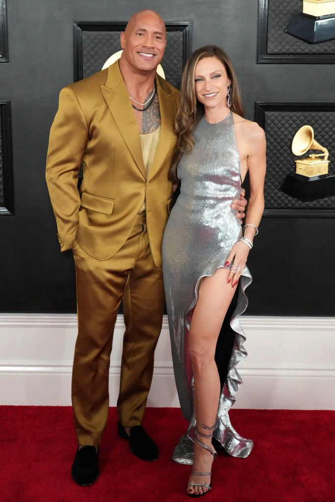 The Rock and Lauren on a red carpet; he&#x27;s in a shiny suit with no tie, and she&#x27;s in a metallic, shimmering, asymmetrical dress with a high slit. Lauren, in heels, comes up to his ear