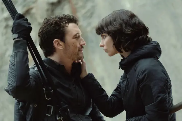 Two people in dark outfits gaze intensely at each other, with one holding climbing gear