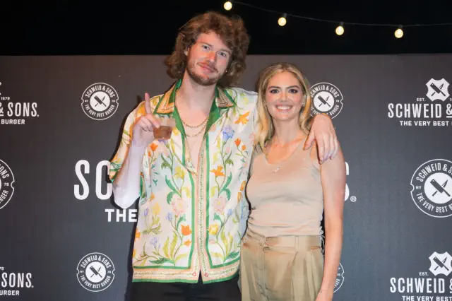 Gravy and Kate pose together; he wears a floral shirt and she&#x27;s in neutral-toned top and pants. They stand in front of a Schweid &amp;amp; Sons backdrop. She comes up to his jaw