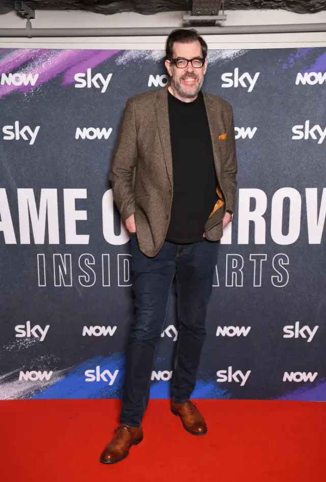 Richard standing on a red carpet, wearing a blazer, shirt, and jeans, smiling