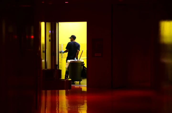 A janitor in uniform works in a dimly lit corridor, pushing a cleaning cart into an elevator