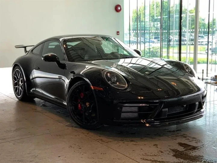 A sleek black Porsche 911 Carrera S is parked indoors, framed perfectly by the large windows in the background. The Carrera S for sale stands out with its elegant design and powerful stance, inviting admiration from every angle.