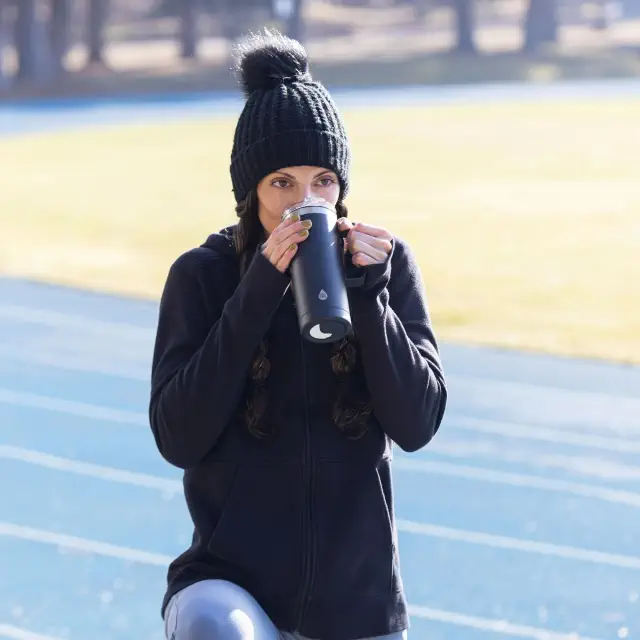 Woman in a knit hat and hoodie drinks from a travel mug while on a track or field, suggesting a sporty and casual outdoor setting