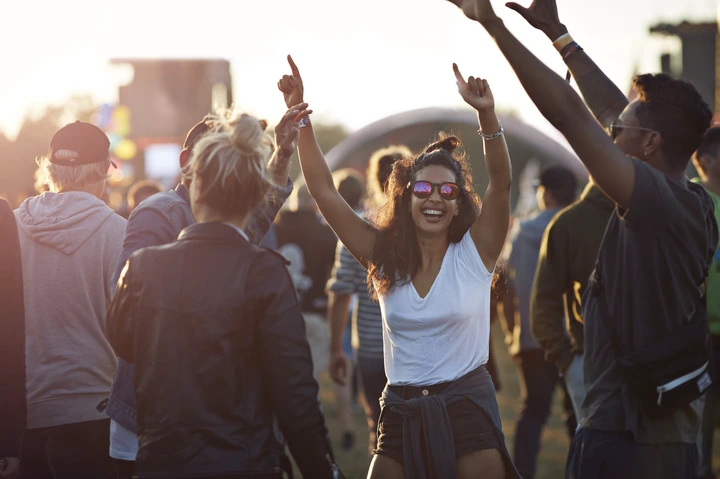 People enjoying an outdoor music festival, dancing and smiling, with one woman in sunglasses and a white shirt raising her arms