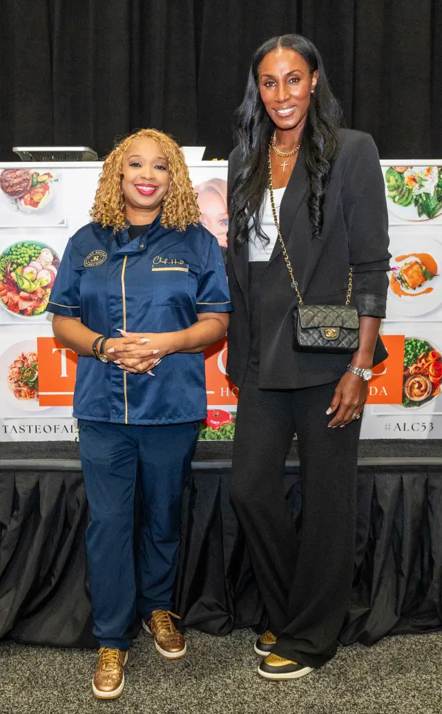 Huda and Lisa stand in front of a culinary backdrop: Huda in a chef outfit, Lisa in elegant attire with a blazer and pants. Huda comes up to Lisa&#x27;s shoulder