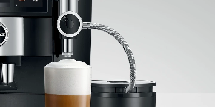 Close-up of a coffee machine frothing milk into a glass of coffee, with a metal steam wand and a milk container on the side, completing the ultimate coffee station ideal for any enthusiast seeking JURA Accessories.
