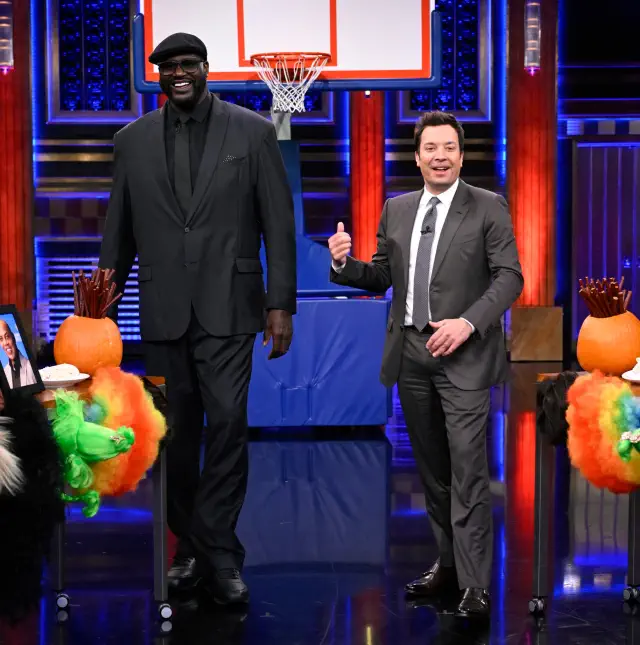 Shaq and Jimmy in suits on a talk show set, smiling and posing near basketball-themed decorations. Jimmy barely comes up to Shaq&#x27;s shoulder