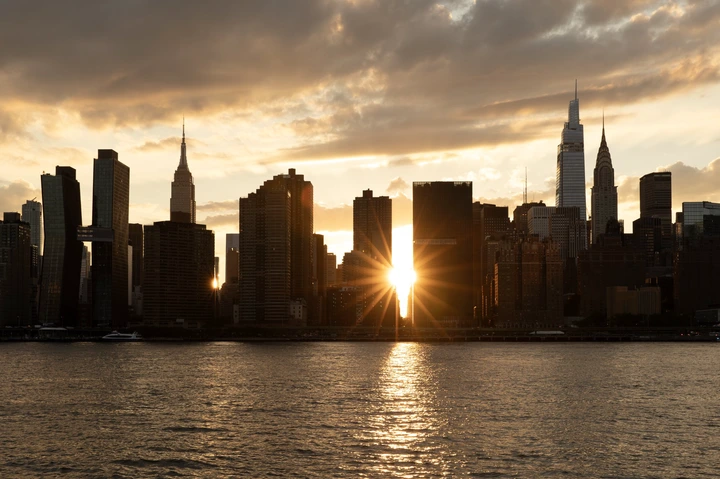 Sunset over a city skyline with skyscrapers silhouetted against the sky, a bright sun flare shining between the buildings