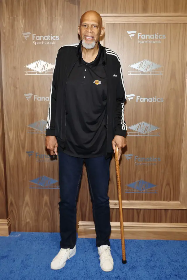Kareem on a red carpet with a casual jacket, dark shirt, and jeans, holding a cane