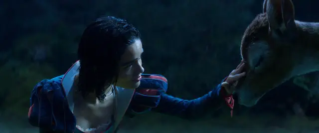 A person in a fairytale-style outfit gently touches the nose of a deer in a dark, forested setting