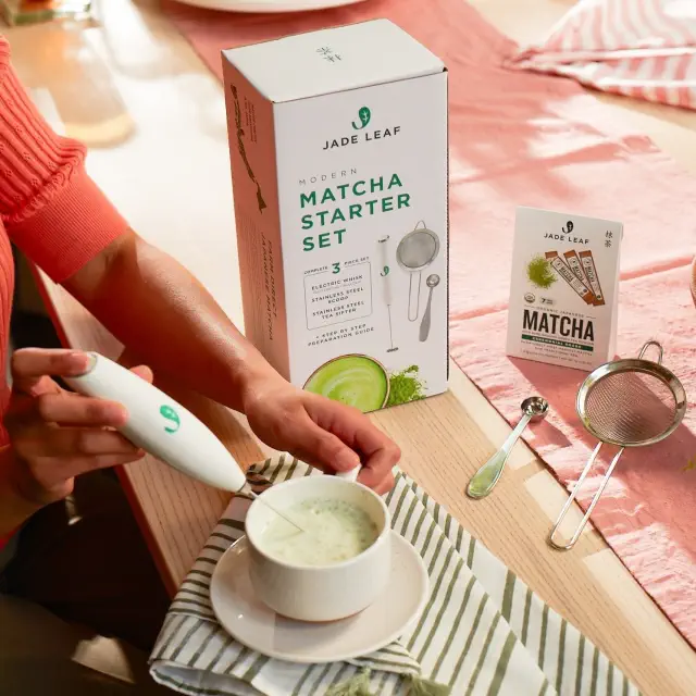 Person using a milk frother with a matcha latte; Jade Leaf Matcha Tea Starter Set and ingredients displayed on a table
