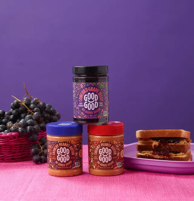 Jars of sugar-free grape and peanut spreads are stacked beside a basket of grapes and a peanut butter and jelly sandwich on a plate