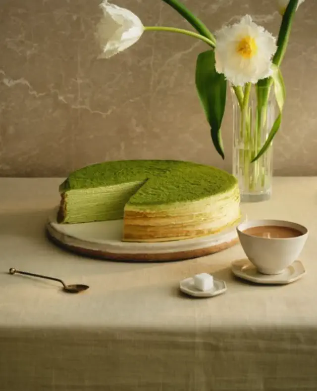 Matcha layer cake with one slice missing, displayed on a table. Beside it are a cup of tea, sugar cubes, and a vase with flowers