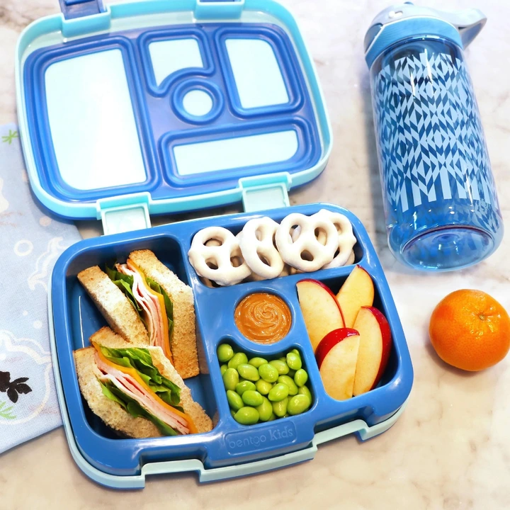 Bento lunchbox with sandwich, apple slices, yogurt pretzels, edamame, dip compartment, next to a water bottle and an orange on a marble counter