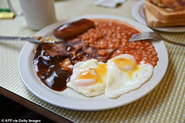 A traditional English breakfast featuring eggs, baked beans, crispy bacon, sausage, and rich gravy is presented on a table at the Regency Cafe in London (stock image).