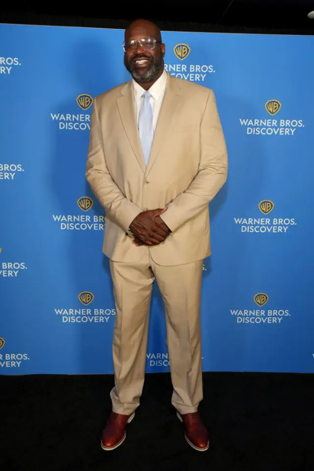 Shaq in a neutral suit and tie on a red carpet, smiling, stands before a Warner Bros. Discovery backdrop