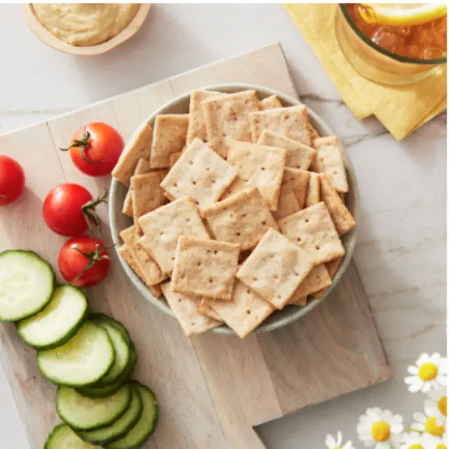 A bowl of square crackers surrounded by cherry tomatoes, sliced cucumber, and a bowl of dip; next to a drink, arranged on a wooden board