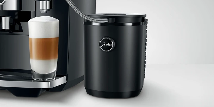 A Jura coffee machine sits proudly at the ultimate coffee station, accompanied by a sleek black milk cooler featuring the Jura logo. A glass of beautifully layered coffee completes this exquisite setup, showcasing the essence of quality with JURA accessories.