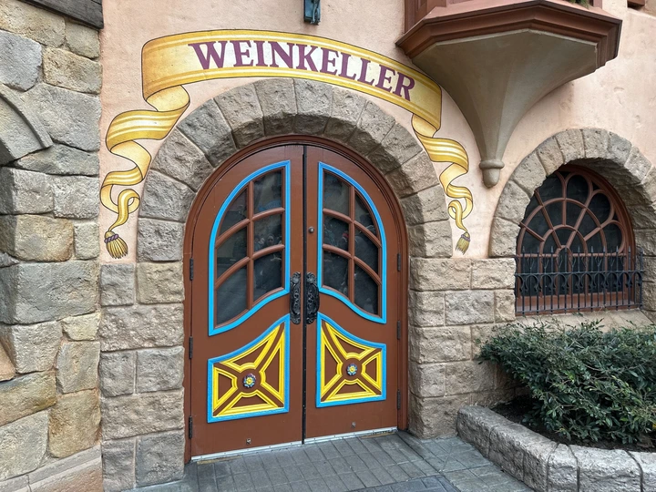 Arched wooden door with colorful geometric designs and "Weinkeller" sign above. Stone wall surrounds the entrance.