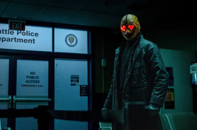 Masked figure with heart-shaped glowing eyes stands in a dimly lit hallway in front of police department doors