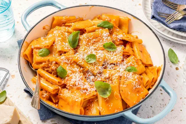 Pot of Paccheri With Quick Sausage Ragu With a Spoon, and in the Surroundings, a Glass, a Block of Parmesan, a Stack of Plates, and Basil Leaves
