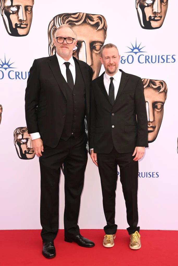 Greg and Alex in suits on a red carpet, Alex wearing gold sneakers, in front of a &quot;Cruises&quot; backdrop with BAFTA mask designs. Alex comes up to Greg&#x27;s ear