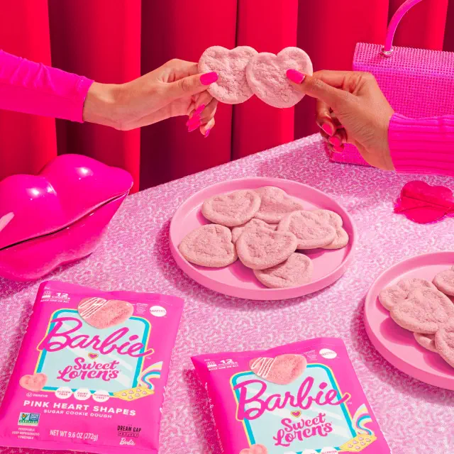 Two hands hold a heart-shaped pink cookie over a table with pink Barbie-themed cookie packaging and accessories, conveying a playful shopping theme