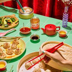 Various dumplings and sauces are set on a dining table with steaming pots, a tea set, and a red backdrop. Hands use chopsticks to serve