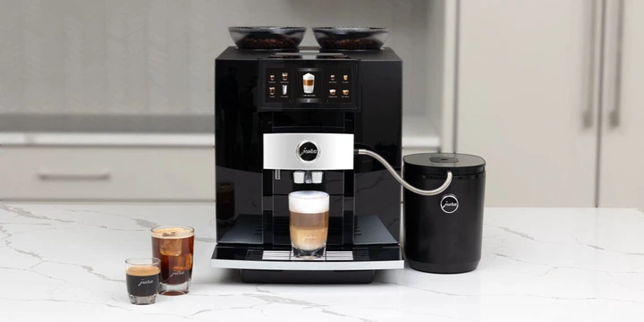 A sleek black JURA espresso machine sits elegantly on a white countertop, surrounded by exquisite coffee station accessories, with two perfectly brewed coffee drinks in cups beside it.