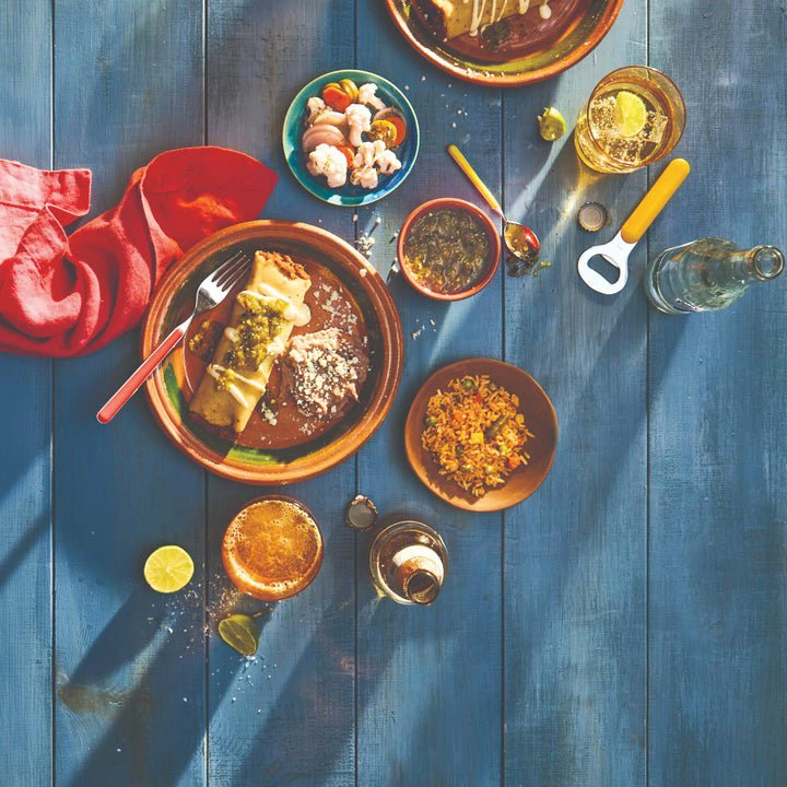 Table setting with two plates of enchiladas, rice, beans, shrimp cocktail, and drinks, on a wooden surface with sunlight