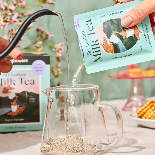 Hands pouring Sri Lankan milk tea mix and hot water into a glass mug on a table set with snacks and flowers. Package in the background
