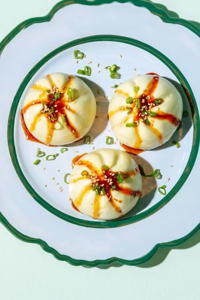 Three round bao buns on a white plate with green trim, topped with green onions, sesame seeds, and sauce drizzles