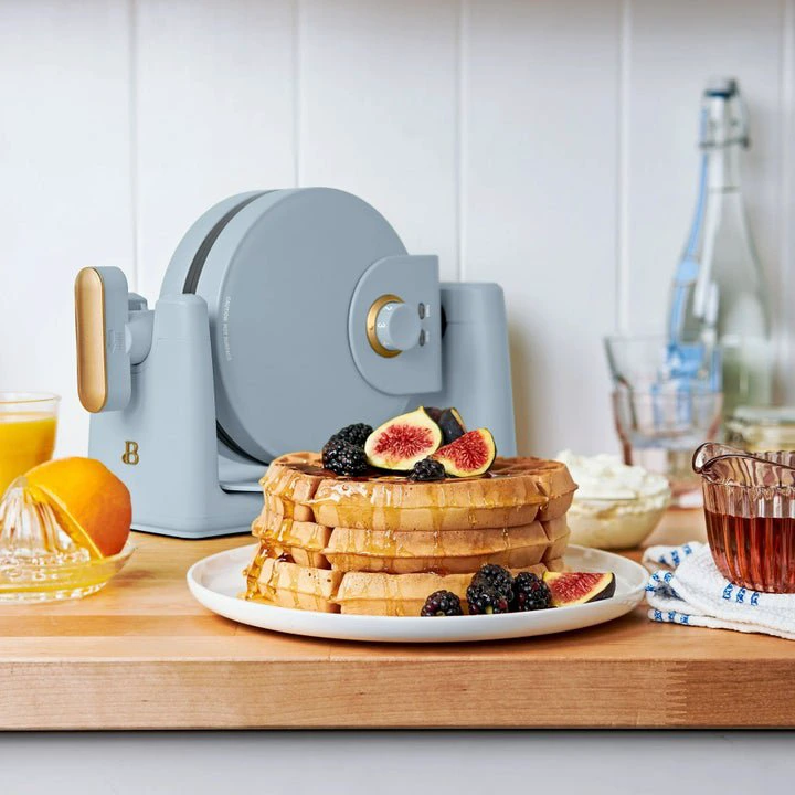 Waffle maker on a kitchen counter with waffles, figs, blackberries, orange juice, cream, and syrup, creating a cozy breakfast scene