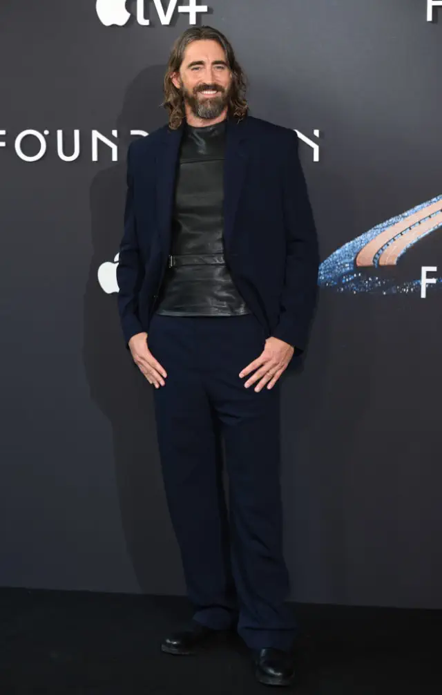 Lee in a dark suit and leather shirt on a red carpet at an Apple TV+ event backdrop