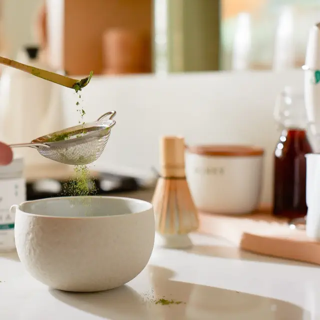 Person sifting matcha powder into a bowl on a kitchen counter; tea accessories and container visible